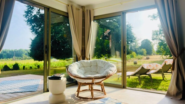A room with a large glass door leading to a garden, a white cushioned chair, a small white table, and a patterned rug.