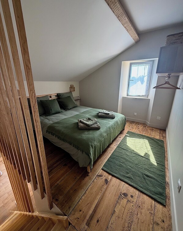 Ein Schlafzimmer mit schrägverdeckter Decke, Holzboden, einem Bett mit grüner Bettwäsche, einem Fenster mit Blick und einem an der Wand hängenden Kleiderhaken.