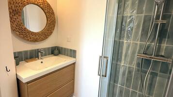 Un bany amb un mirall rodó, un lavabo, una dutxa amb porta de vidre i un armari de fusta.