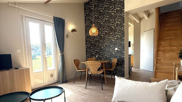 Quarto de hotel moderno com uma cama grande, área de jantar com cadeiras de vime, uma pequena mesa e vista para o exterior por uma porta deslizante.