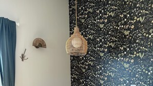 Uma sala de jantar com uma mesa de madeira redonda, cadeiras de vime, um lustre pendurado e uma parede com padrão floral no papel de parede.