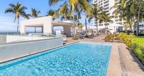Une piscine entourée de chaises longues et de palmiers, le tout bordé d’un bâtiment moderne doté de balcons.