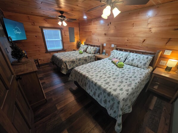 A wooden cabin room with two beds, a ceiling fan, a small plant, and a Disney+ sign. - Country Cabin - Hot Tub - Free Kayaks - Gig Speed Internet - Fire Pit! (Loogootee)