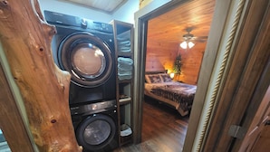 Une buanderie équipée d’un lave-linge et d’un sèche-linge, un plancher en bois, et une chambre avec un lit et un ventilateur de plafond.