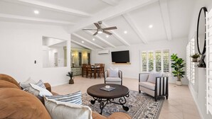 A spacious living room with a high ceiling, a ceiling fan, a dining area with a TV, and a large window with blinds.