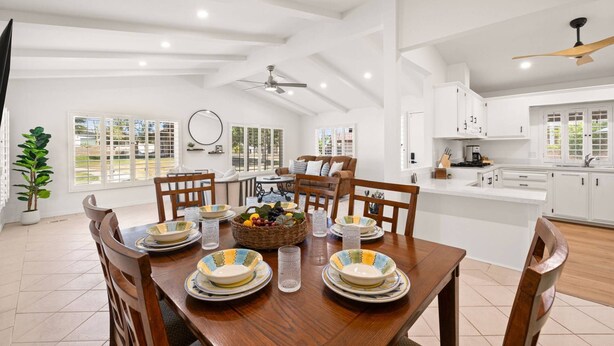 A spacious dining area with a wooden table set for six, surrounded by chairs. A large window provides natural light, and the room features white cabinetry and a ceiling fan.