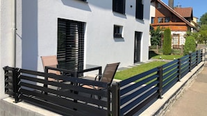 Een wit gebouw met zwarte luiken, een terras met een tafel en stoelen, en een hek van zwart metaal.