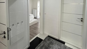 Een gang met een witte deur, een kleedje en een bed op de achtergrond.