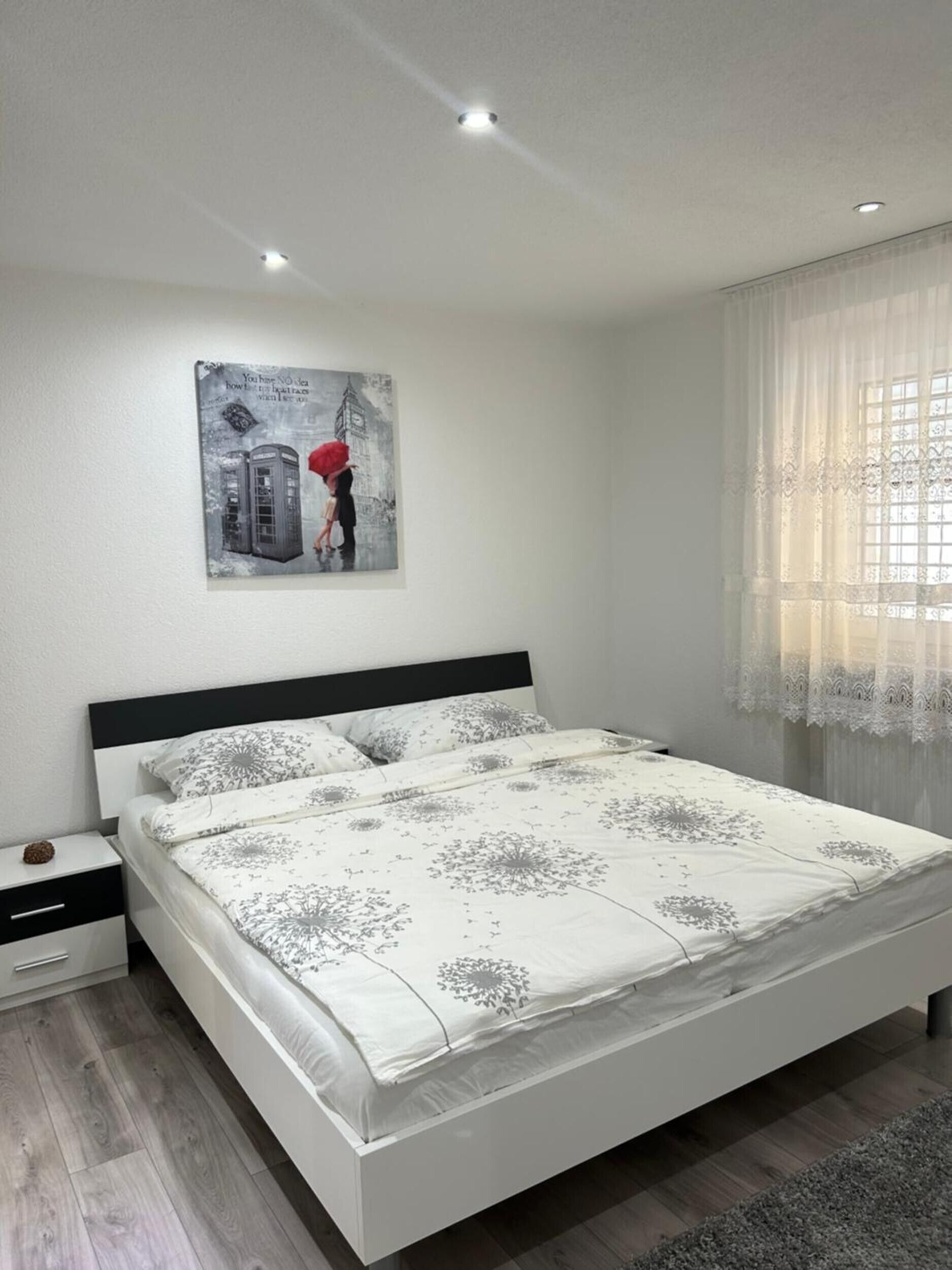 Un dormitorio moderno con cama, mesita de noche, una obra de arte en la pared y una ventana con cortinas traslúcidas.