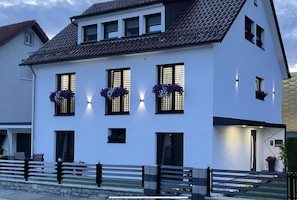 A white building with a tiled roof, multiple windows with black shutters, and a balcony with a railing.