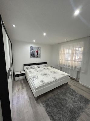 A bedroom with a bed, a nightstand, a wall-mounted light, a window with sheer curtains, and a painting on the wall.