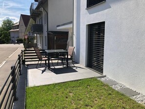 Un bâtiment moderne doté d’un patio comprenant une table et des chaises, un petit jardin et une porte de garage.