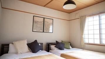 A hotel room with two beds, a wooden ceiling, a large window with curtains, and two framed maps on the wall.