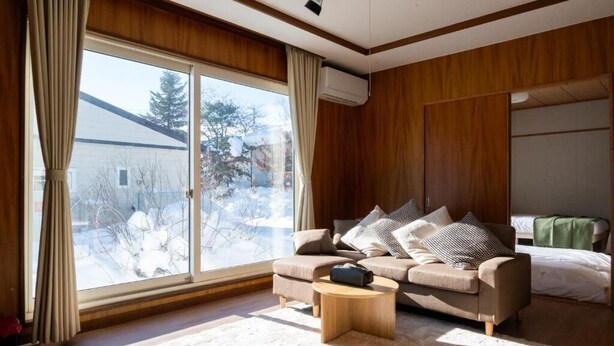 A cozy living room with a sofa, a coffee table, and a large window with curtains. - FURANO RELAX STAY A (Furano)