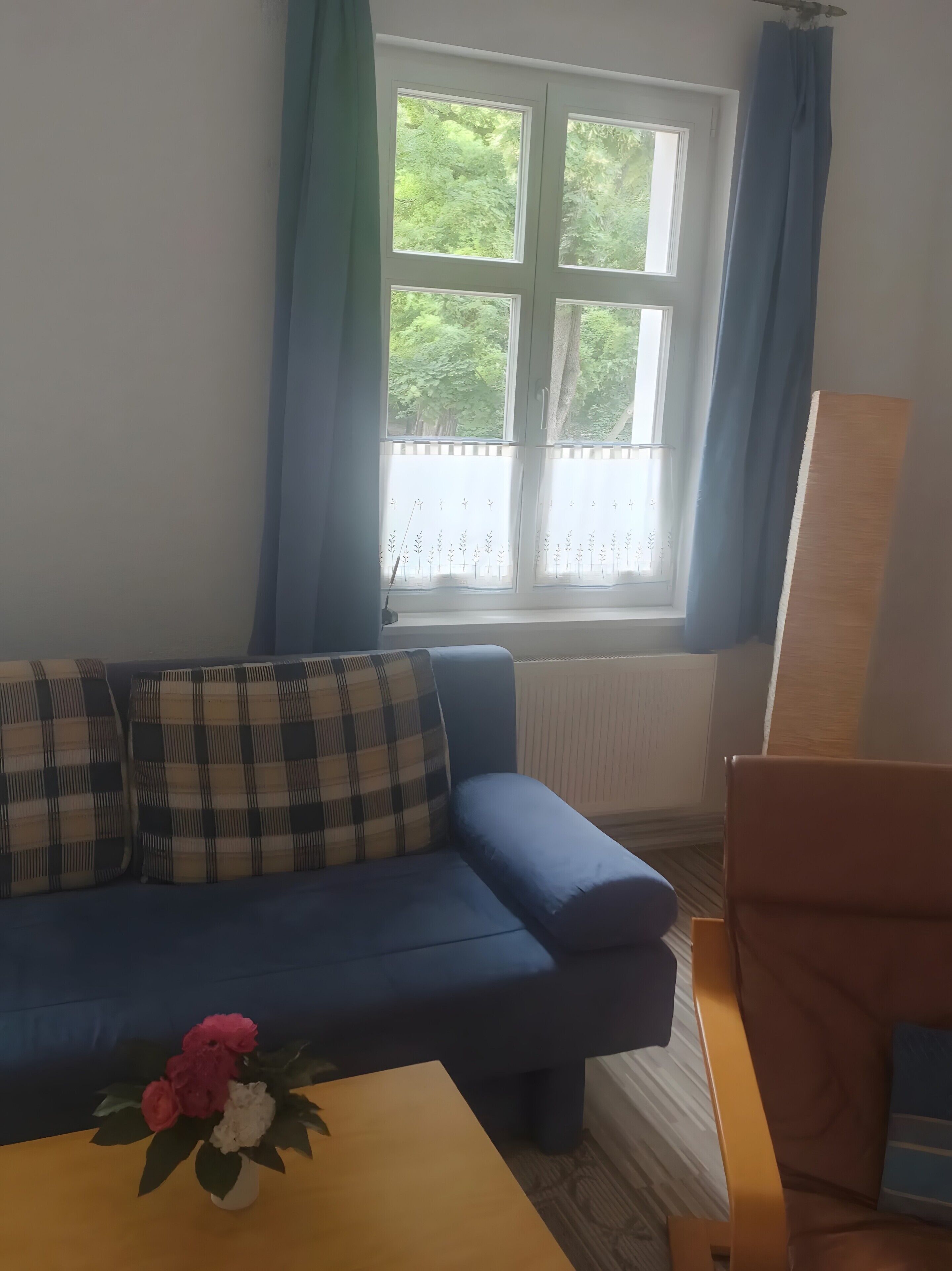 A room with a blue sofa, a wooden table with a vase of flowers, a window with blue curtains, and a radiator.