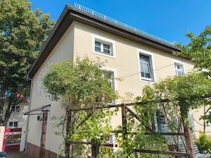 Un edificio de dos pisos con un balcón, una verja de madera y una pérgola con plantas trepadoras.