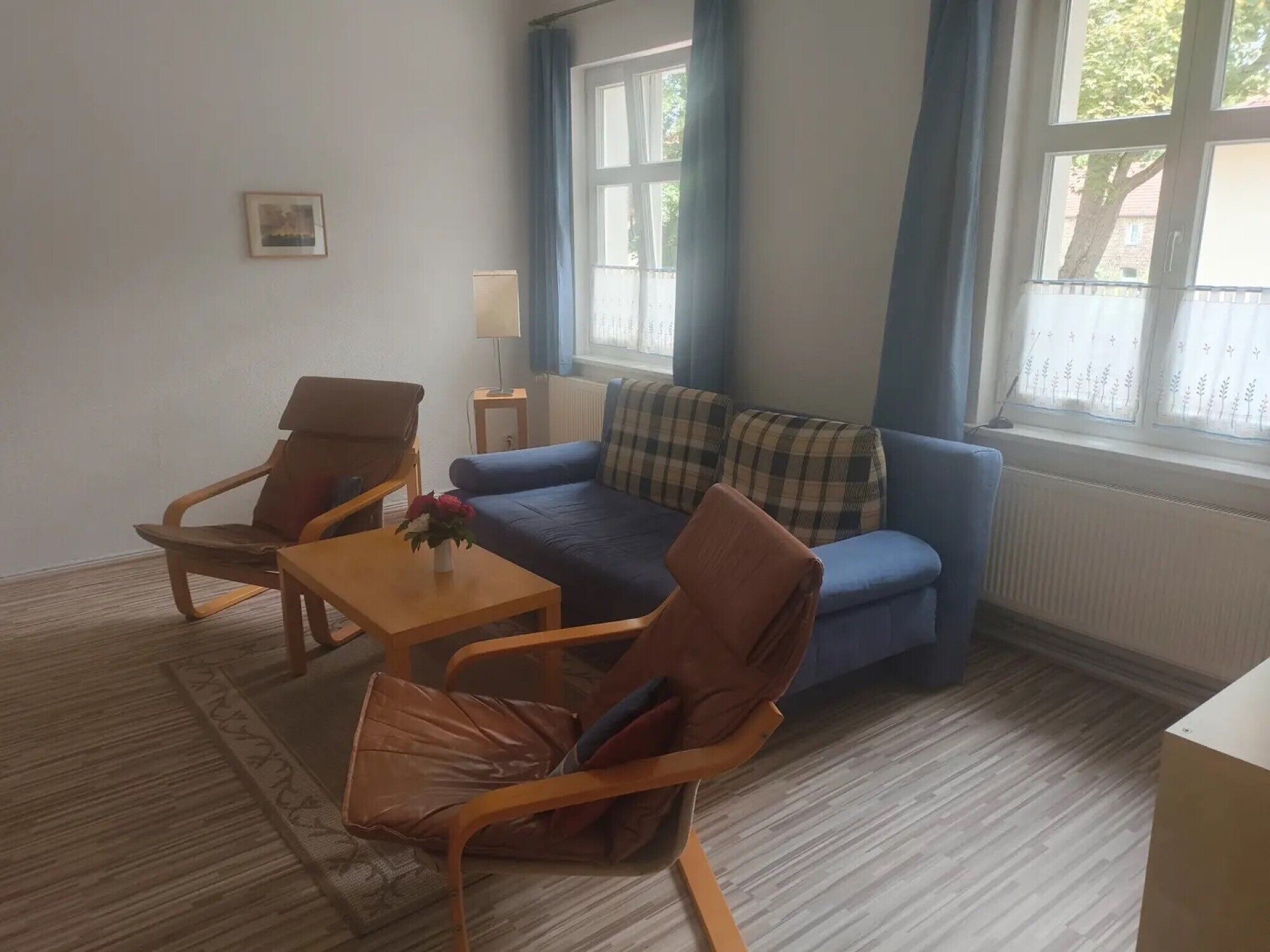 Ein Wohnzimmer mit einem blauen Sofa, zwei Holzsesseln, einem kleinen Tisch mit Blumenvase und einem Fenster mit Vorhängen.