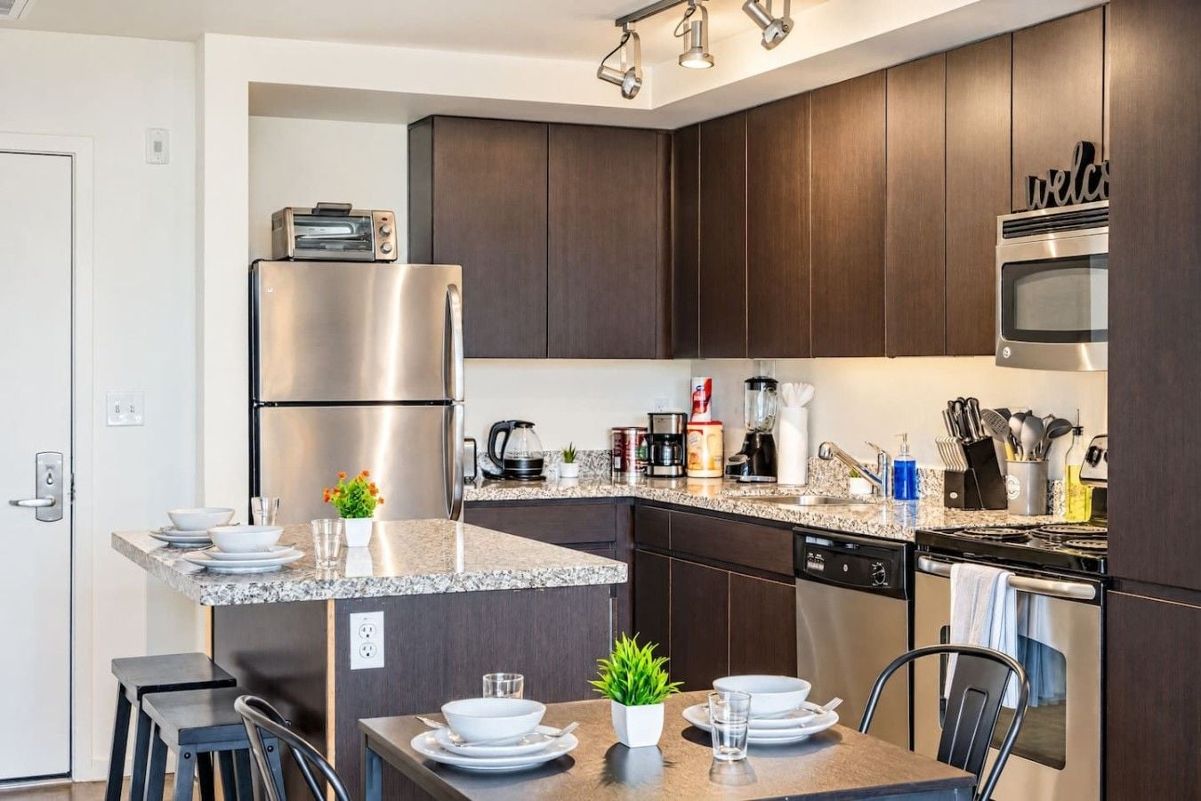 Una cocina moderna con armarios de madera oscura, electrodomésticos de acero inoxidable, una encimera de granito y un comedor con una mesa preparada para dos personas.