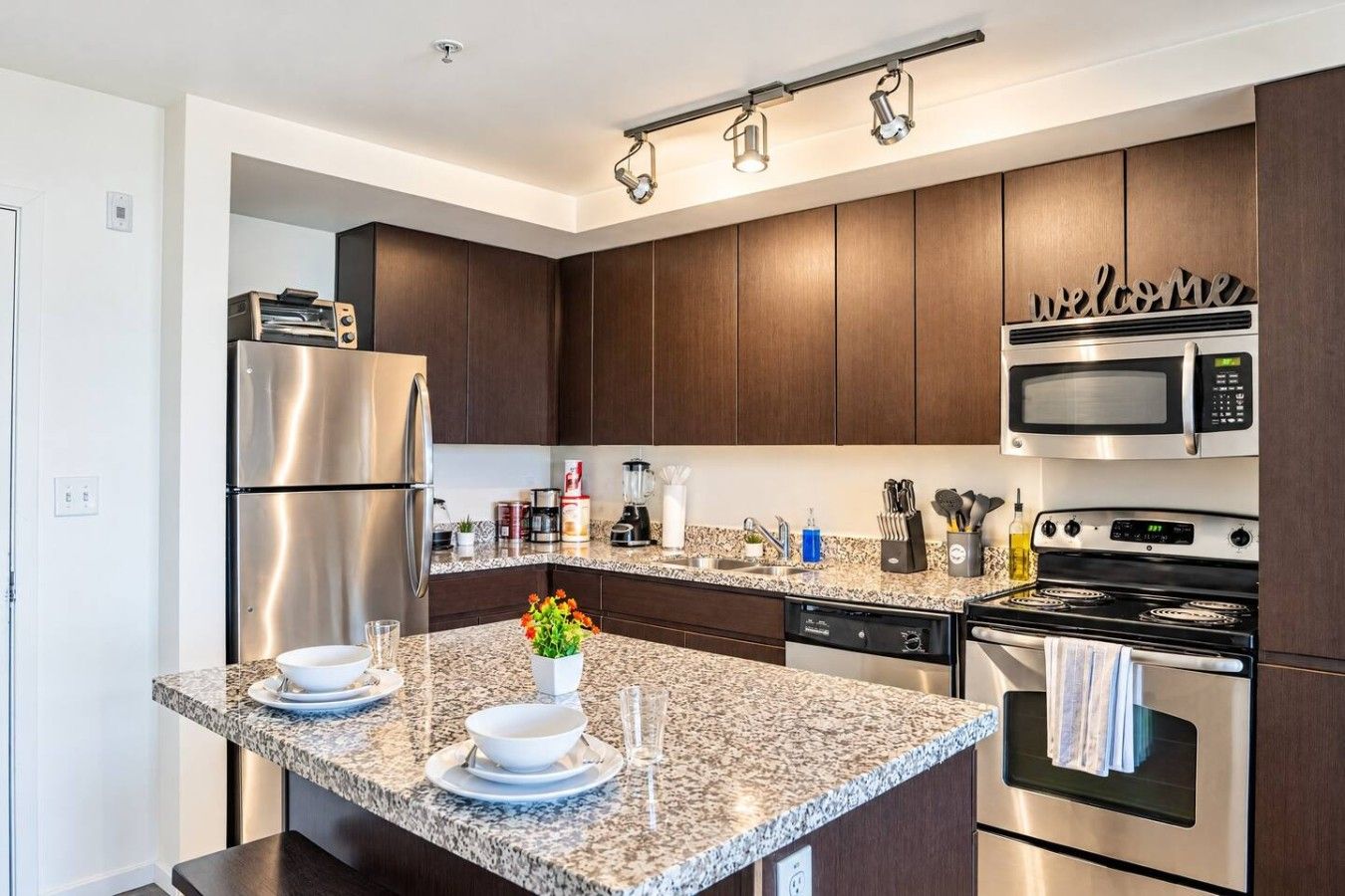 A modern kitchen with dark wood cabinets, stainless steel appliances, a granite countertop, and a welcome sign above the microwave.