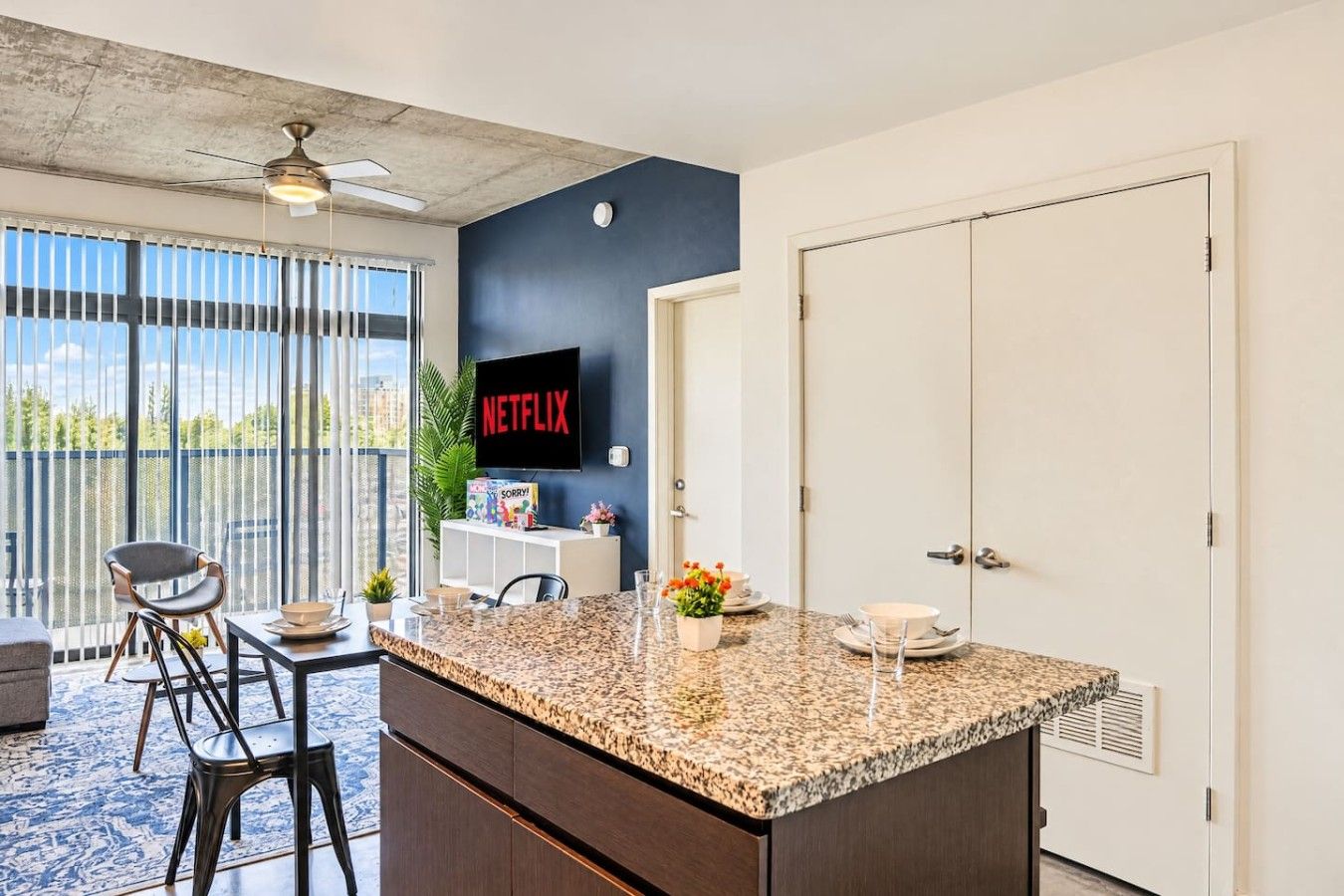 A modern kitchen with a granite countertop, bar stools, a television, and a view through large windows.