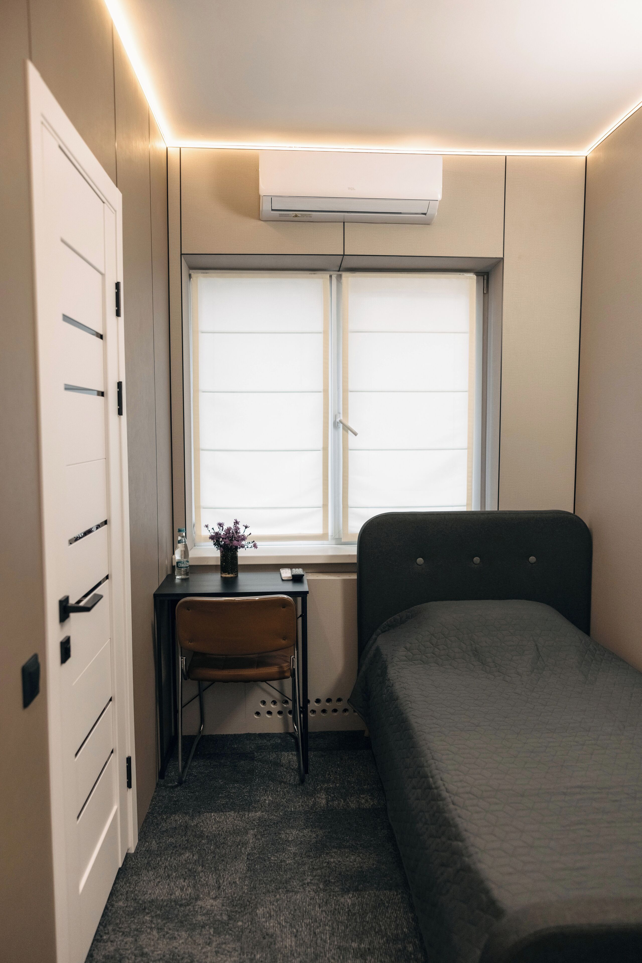 A compact room with a bed, a desk with a chair, a window, and an air conditioning unit.