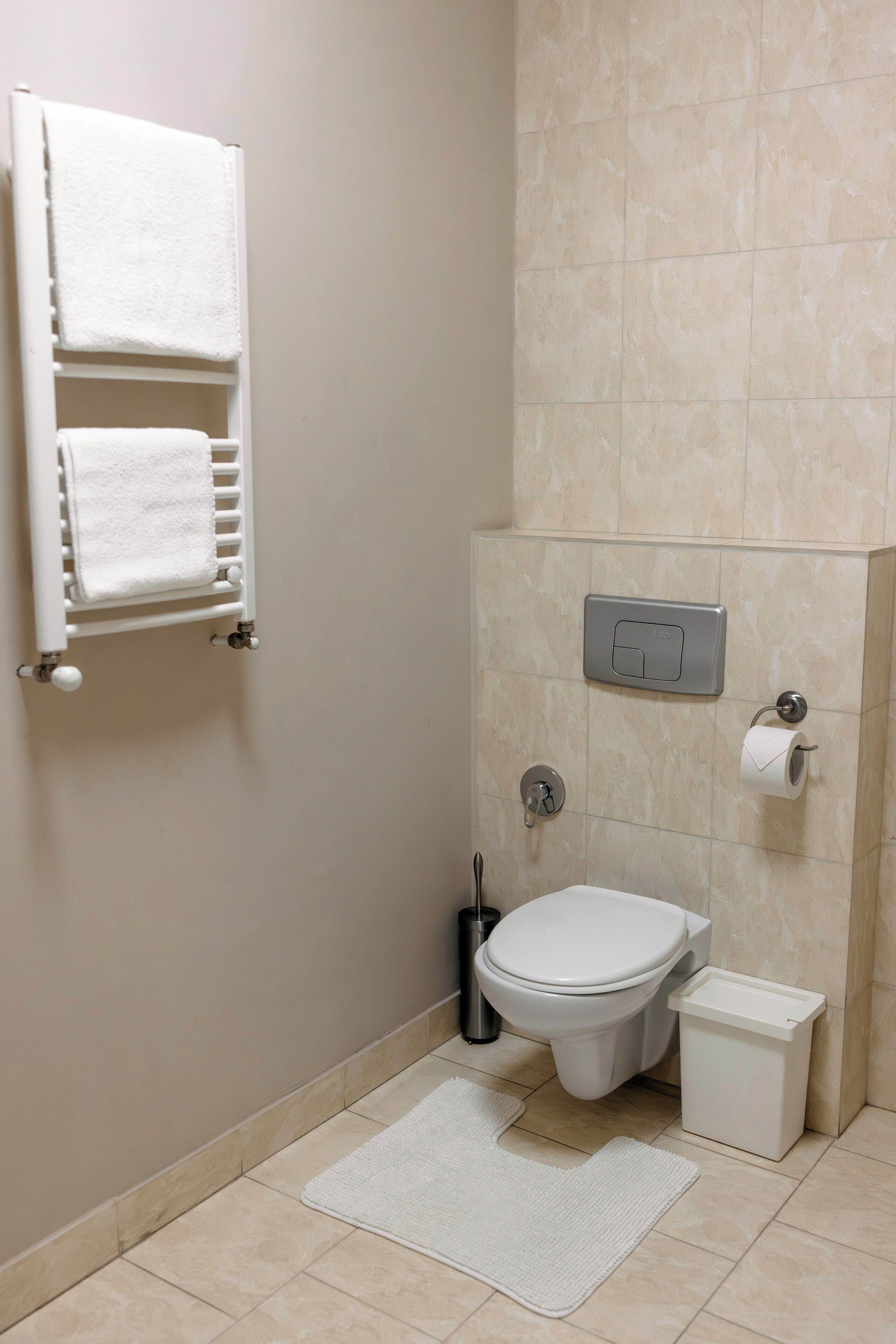 Un baño pequeño y limpio con inodoro, un cubo de basura, toallero con dos toallas y dispensador de papel.