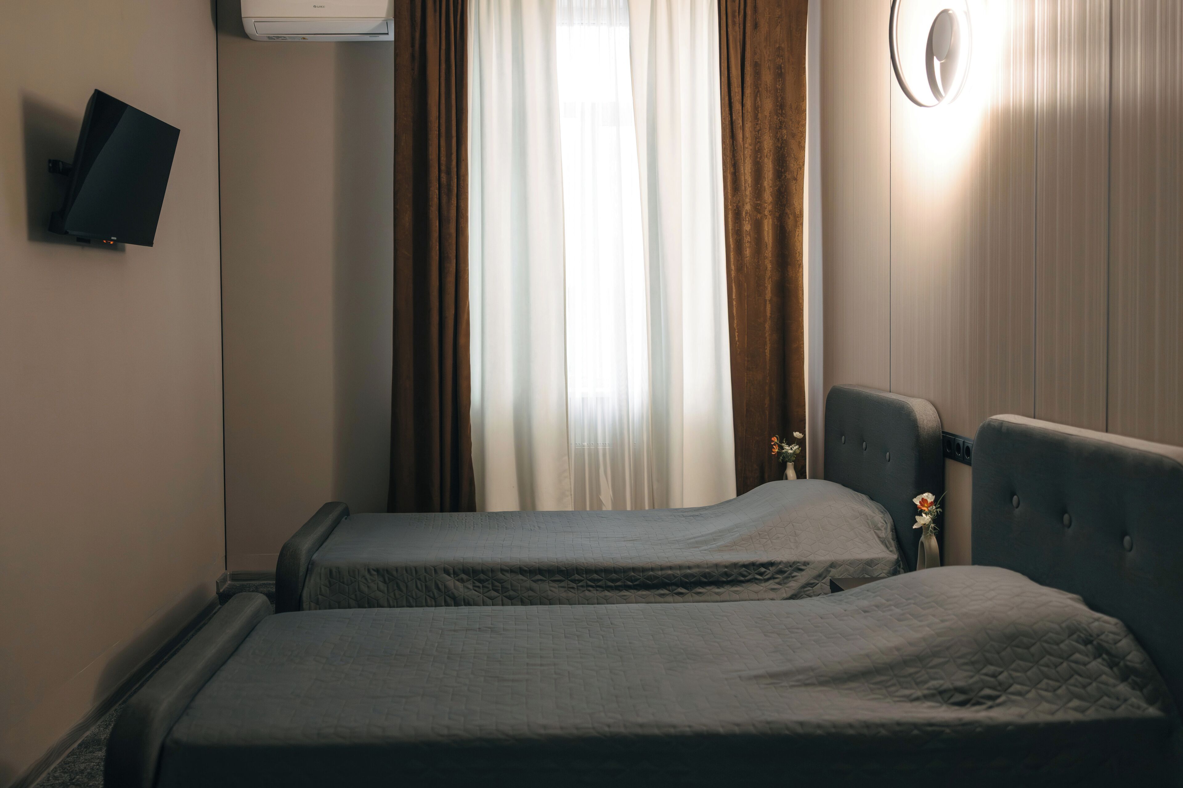 Habitación de hotel con dos camas, un televisor de pantalla plana montado en la pared, una ventana con cortinas y una lámpara fijada a la pared.