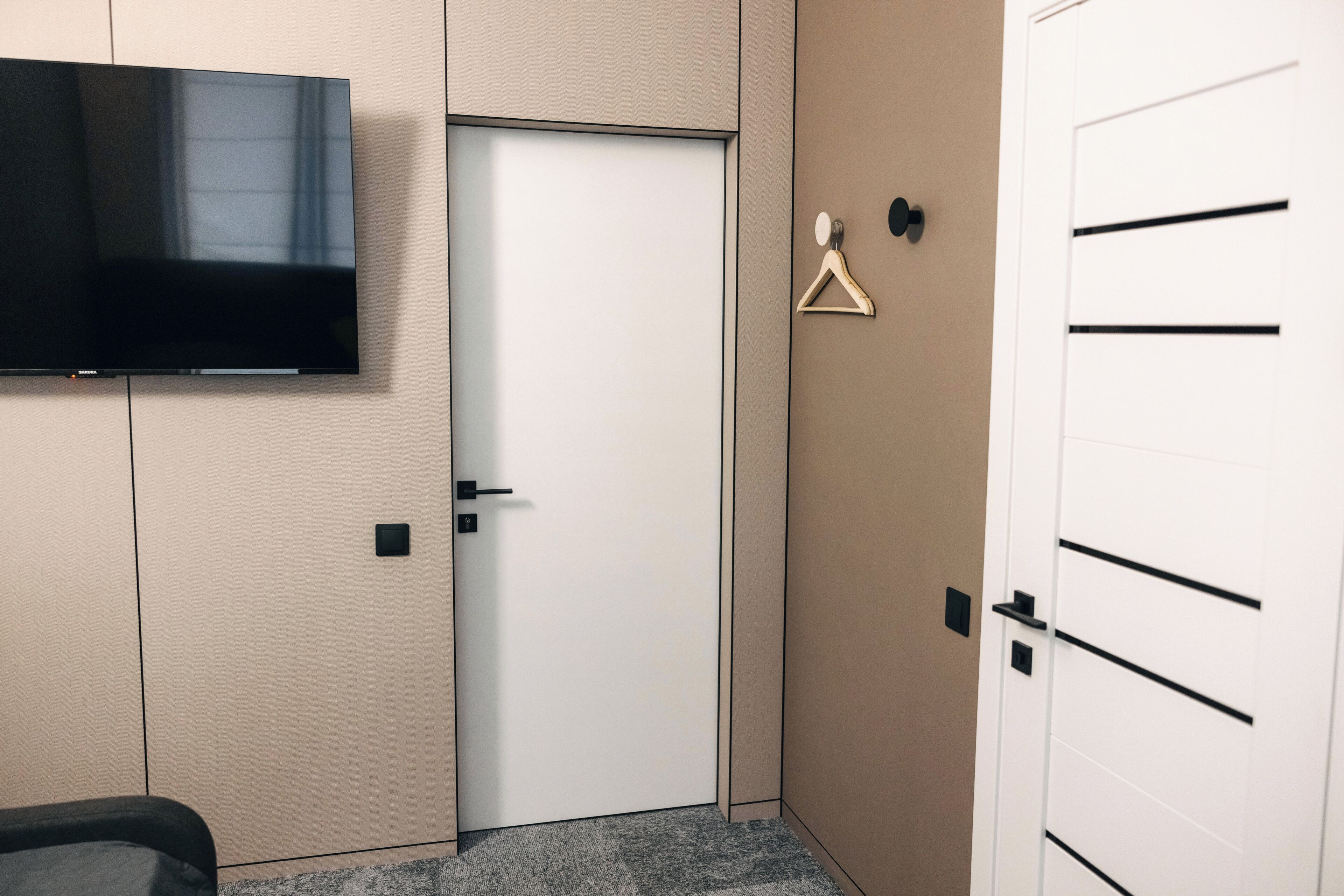 A hotel room with a flat-screen TV mounted on the wall, a closed door, a coat hanger, and a white wardrobe with black handles.