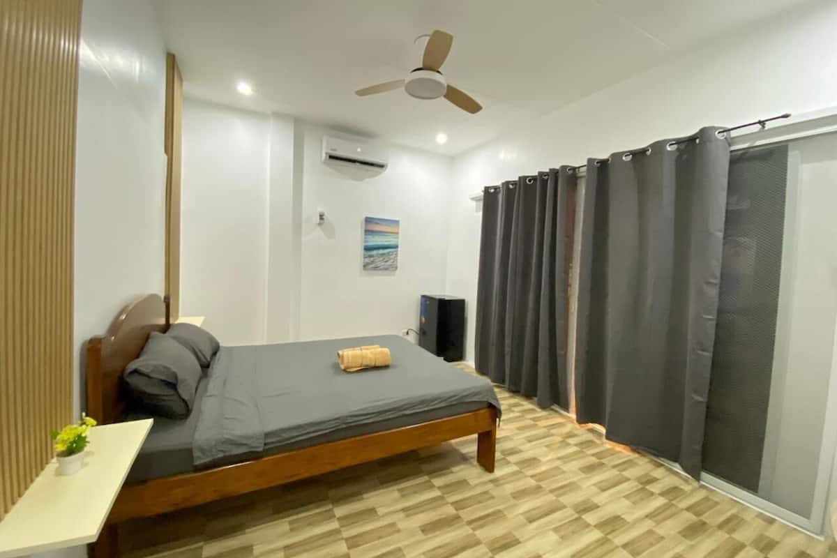 Ein Schlafzimmer mit Bett, Deckenventilator, Klimaanlage, Lautsprecher und einem Fenster mit Vorhängen.
