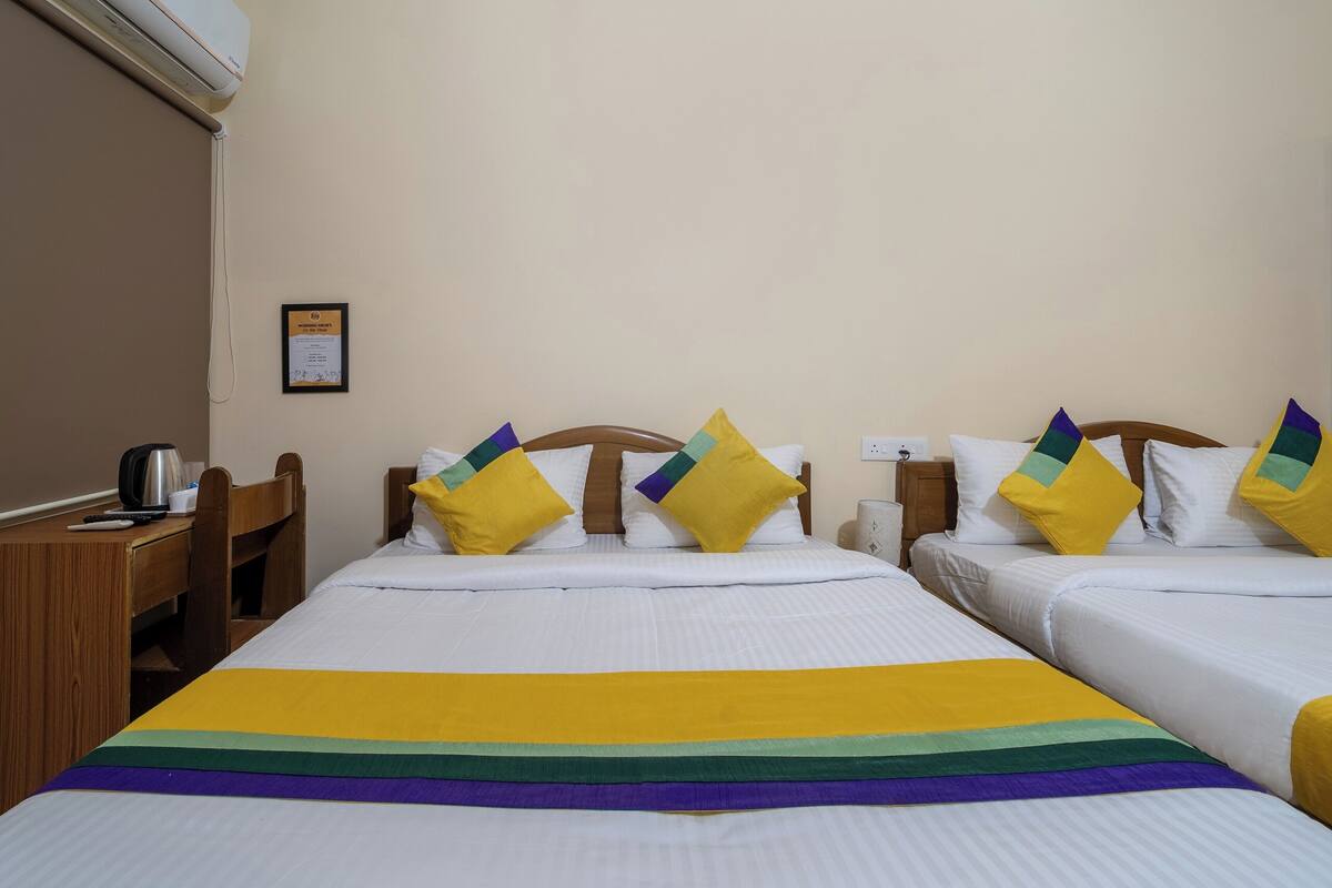 Habitación de hotel con dos camas, cada una con un colchón blanco y una manta colorida. Tiene un cabecero de madera, una mesita de noche con lámpara y un certificado enmarcado en la pared.