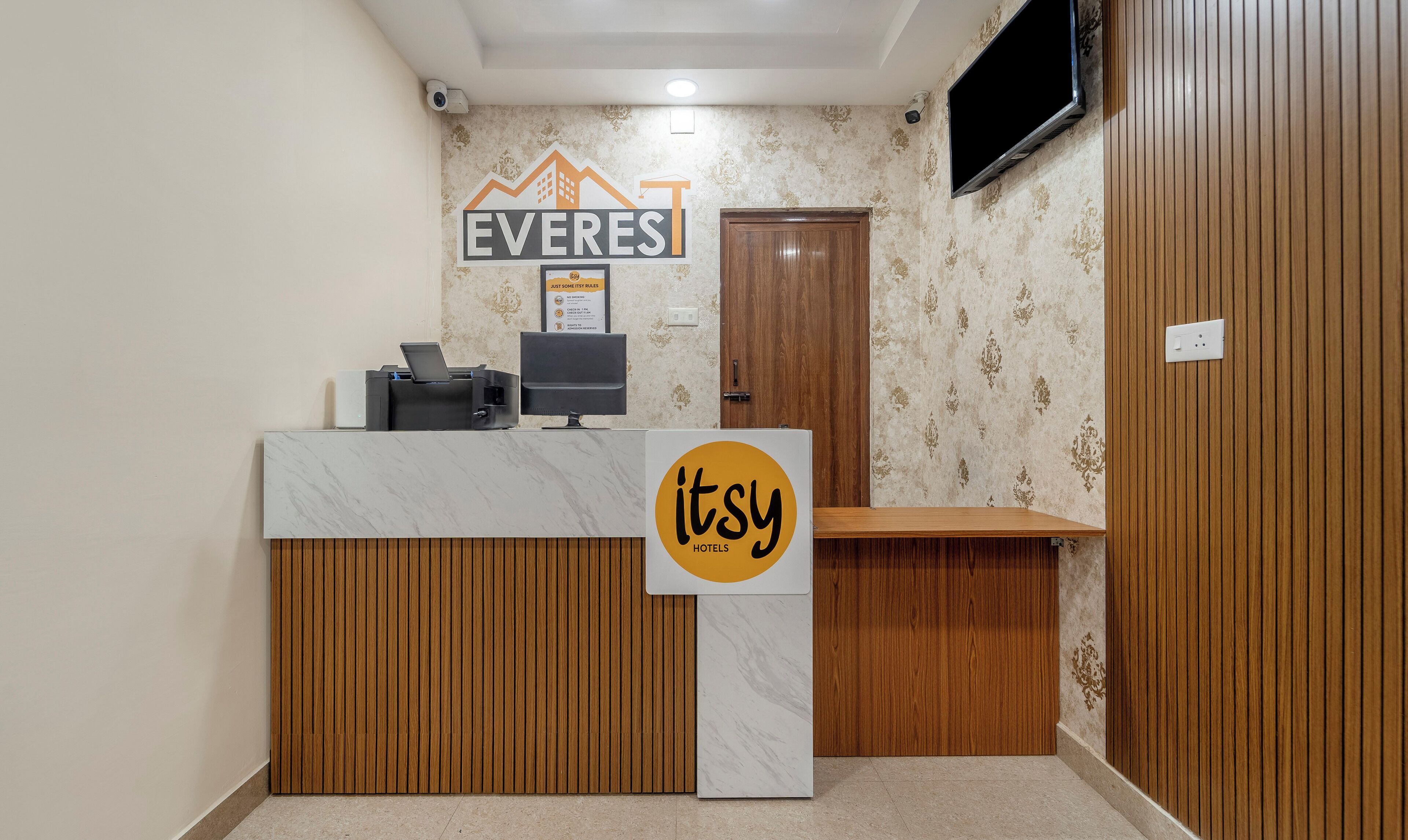 A hotel reception desk with a sign for 'EVERES' and 'etsy hotels' branding, a flat-screen TV mounted on the wall, and a wooden panel design on the walls.