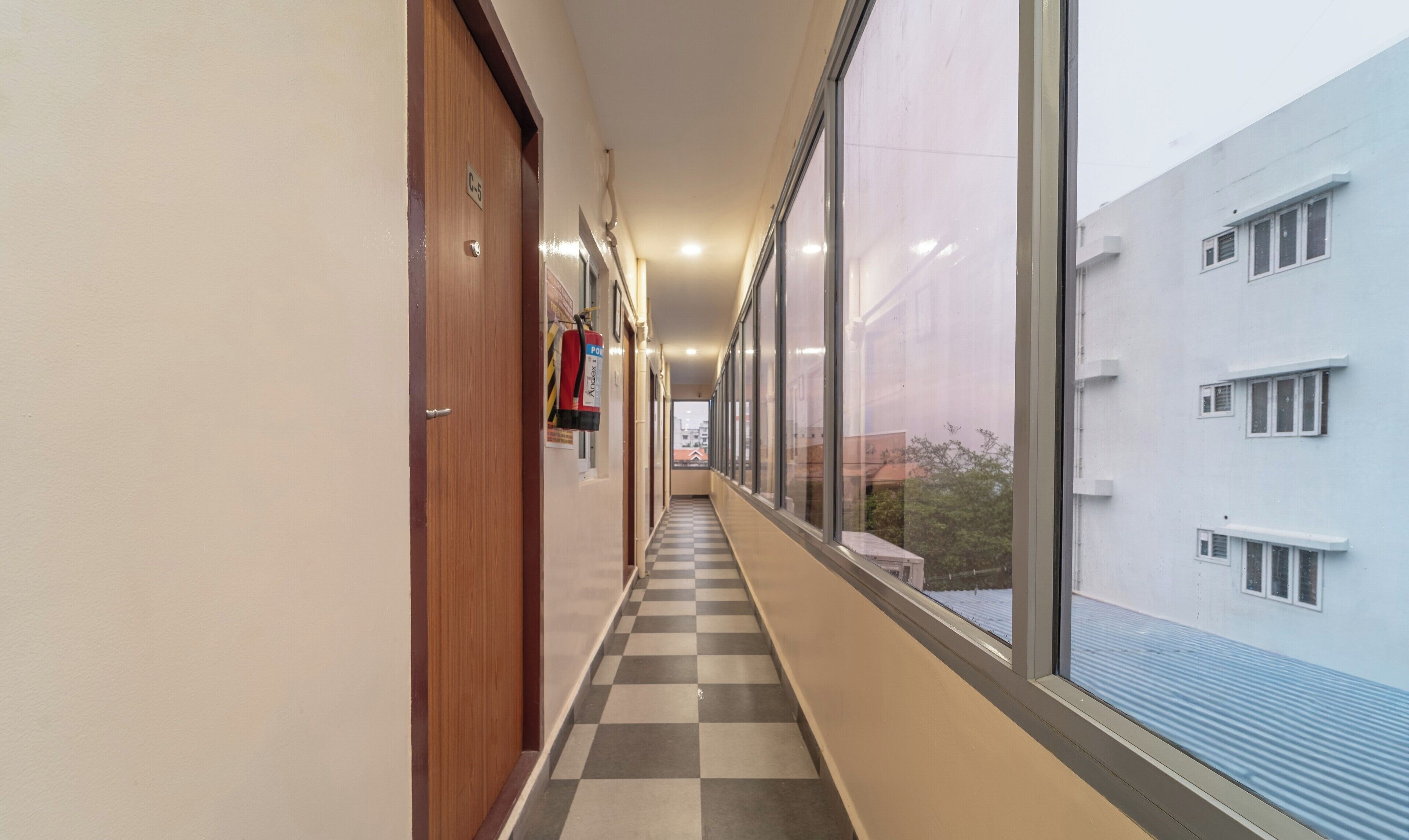 A corridor with checkered flooring, wooden doors, and large windows offering a view of a building with multiple windows.