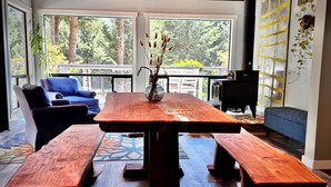 Une pièce spacieuse avec une grande table en bois et des bancs, un canapé bleu et une vue sur les arbres à travers de grandes fenêtres.