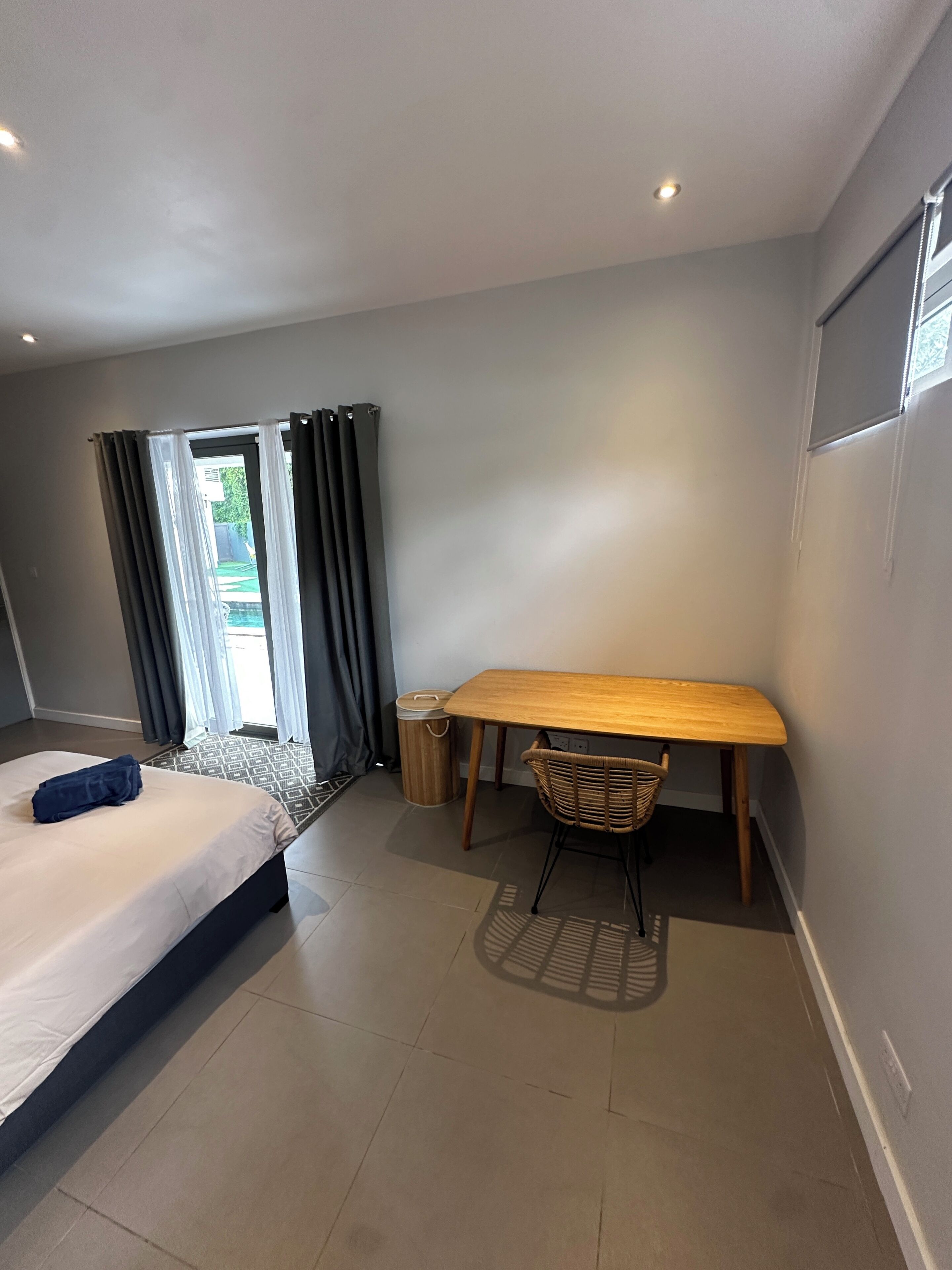 A hotel room with a bed, a wooden desk, a wicker chair, a waste bin, and a view through a window with curtains.