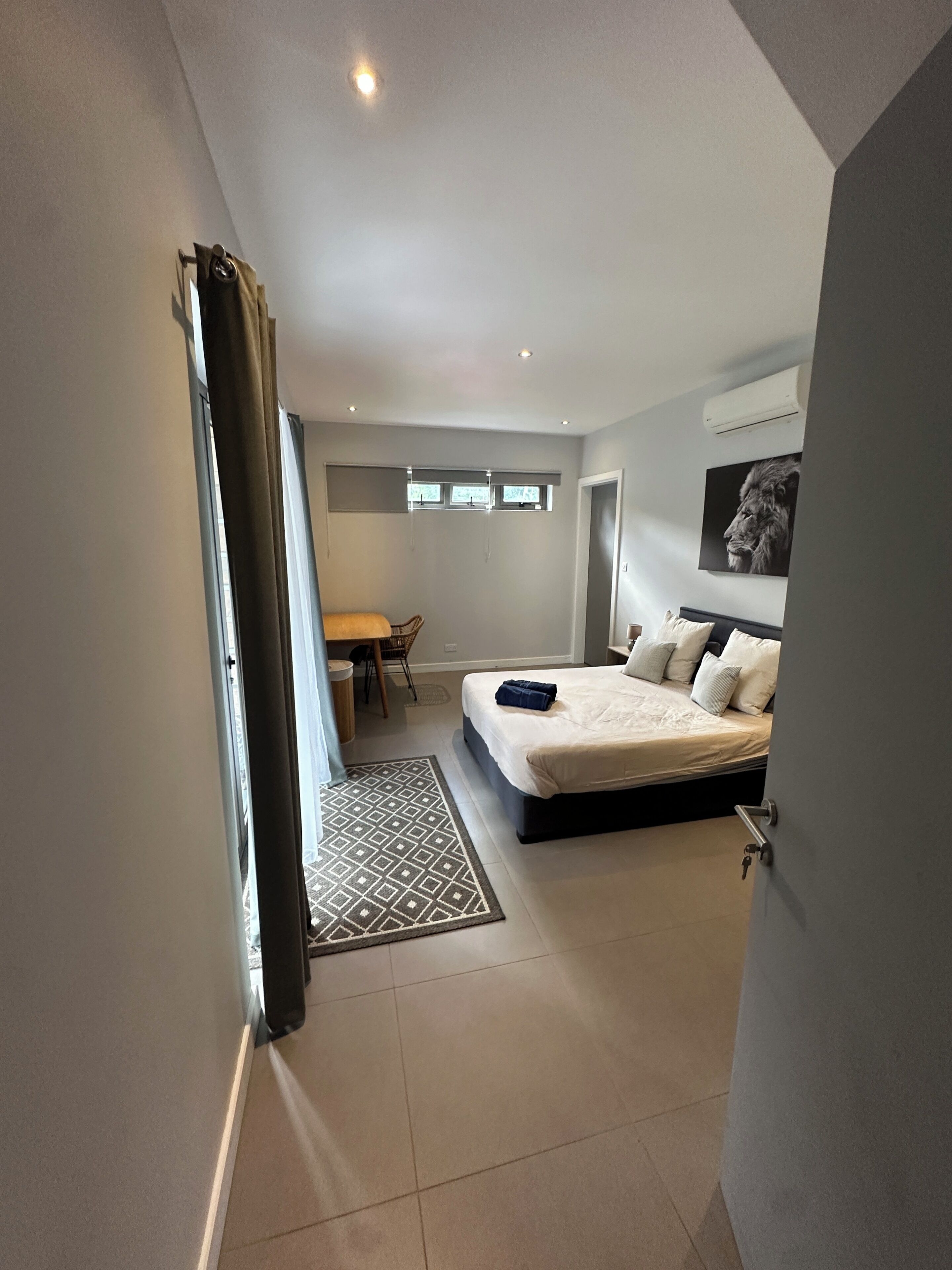 Ein modernes Schlafzimmer mit einem großen Bett, einem Schreibtisch und einem Sessel. Ein Fenster mit Vorhängen, ein gemustertes Teppich und eine wandmontierte Klimaanlage sind ebenfalls vorhanden.
