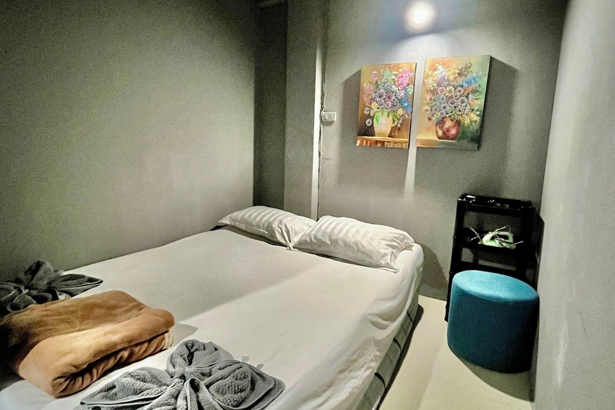 A small, modern room with a single bed, a blue ottoman, and two wall-mounted paintings.