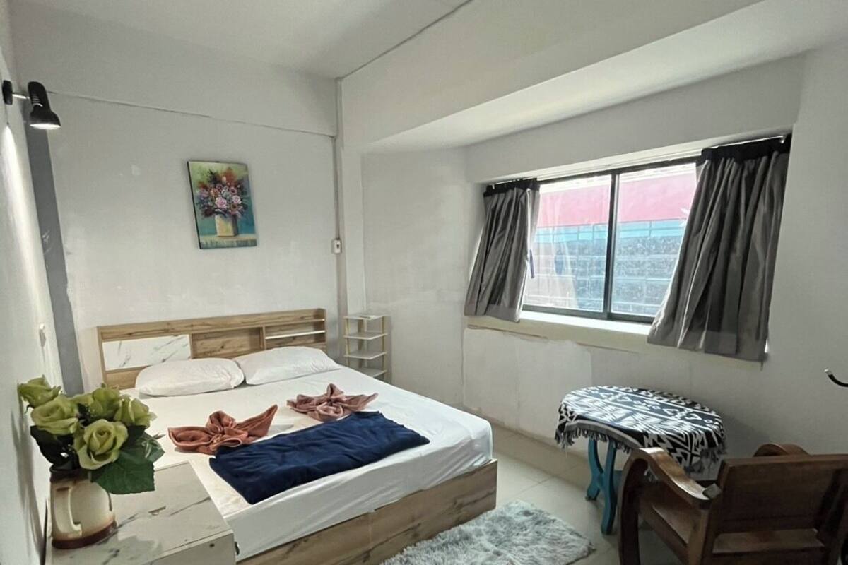 A bedroom with a bed, a desk, a chair, a window with curtains, a small table, and a decorative vase with flowers.