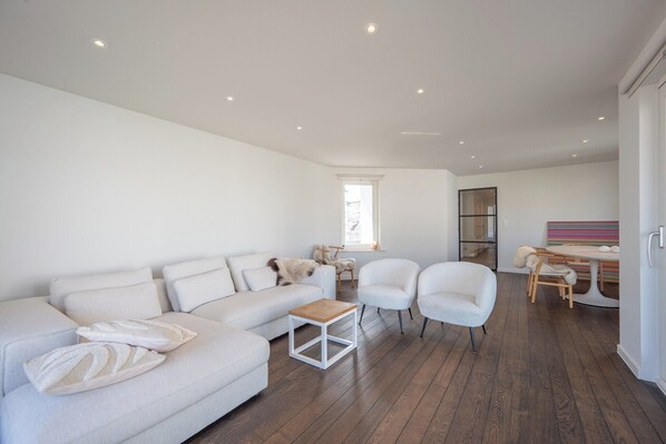 A spacious living room with a white sofa, two armchairs, a wooden coffee table, and a dining area with a colorful striped tablecloth.
