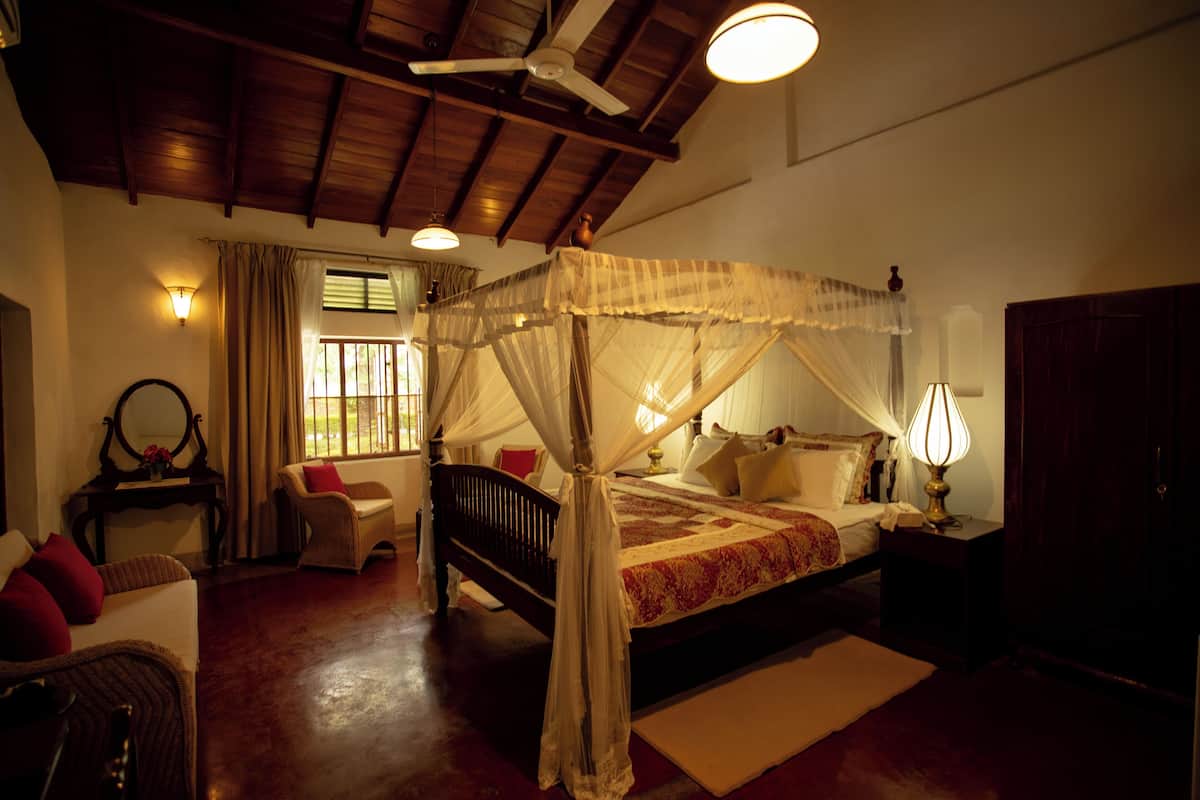 A four-poster canopy bed with a red and white bedspread, a nightstand with a lamp, a wicker chair, and a wooden desk with a mirror.