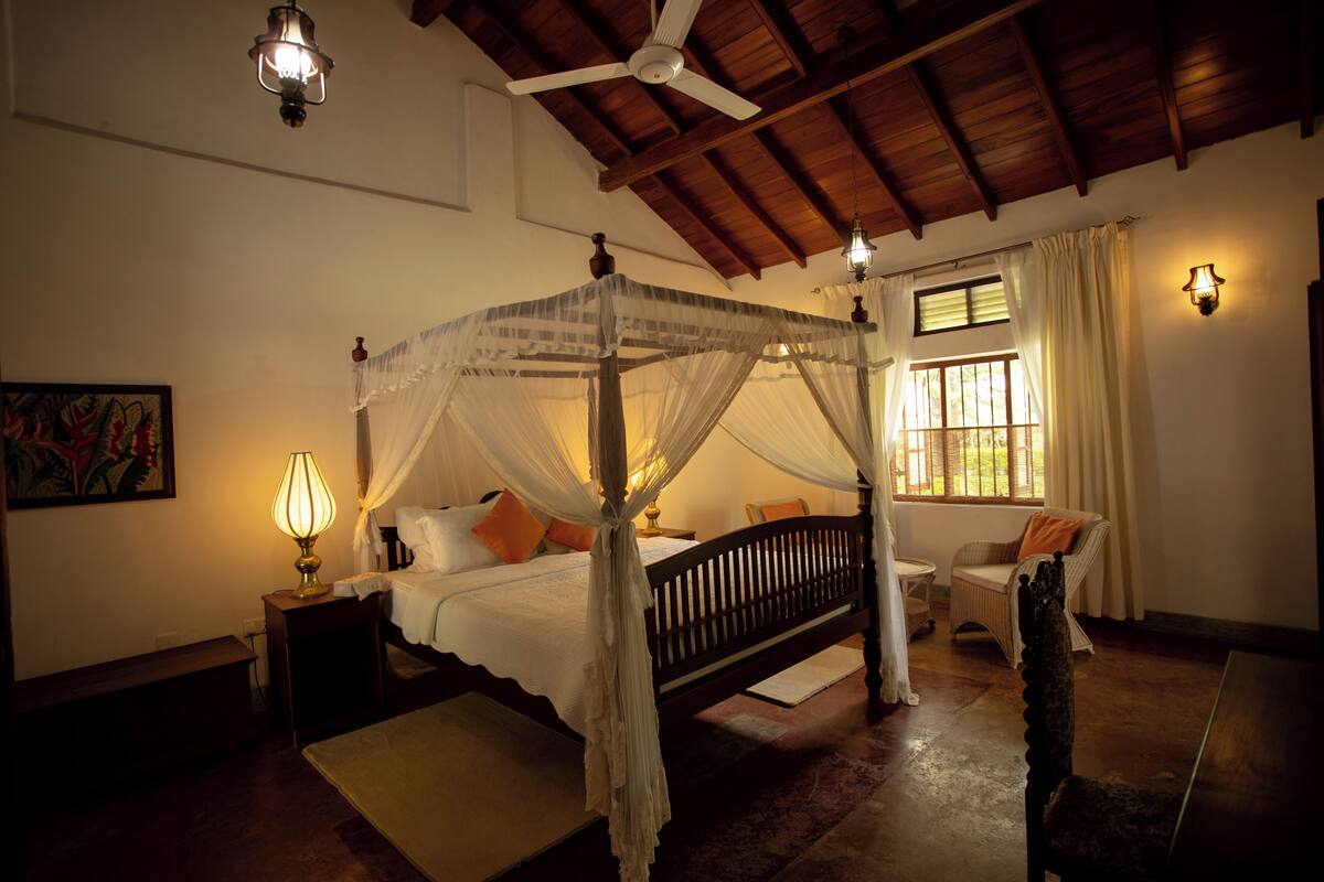 A four-poster bed with a canopy, a bedside table with a lamp, a chair, and a window with curtains.
