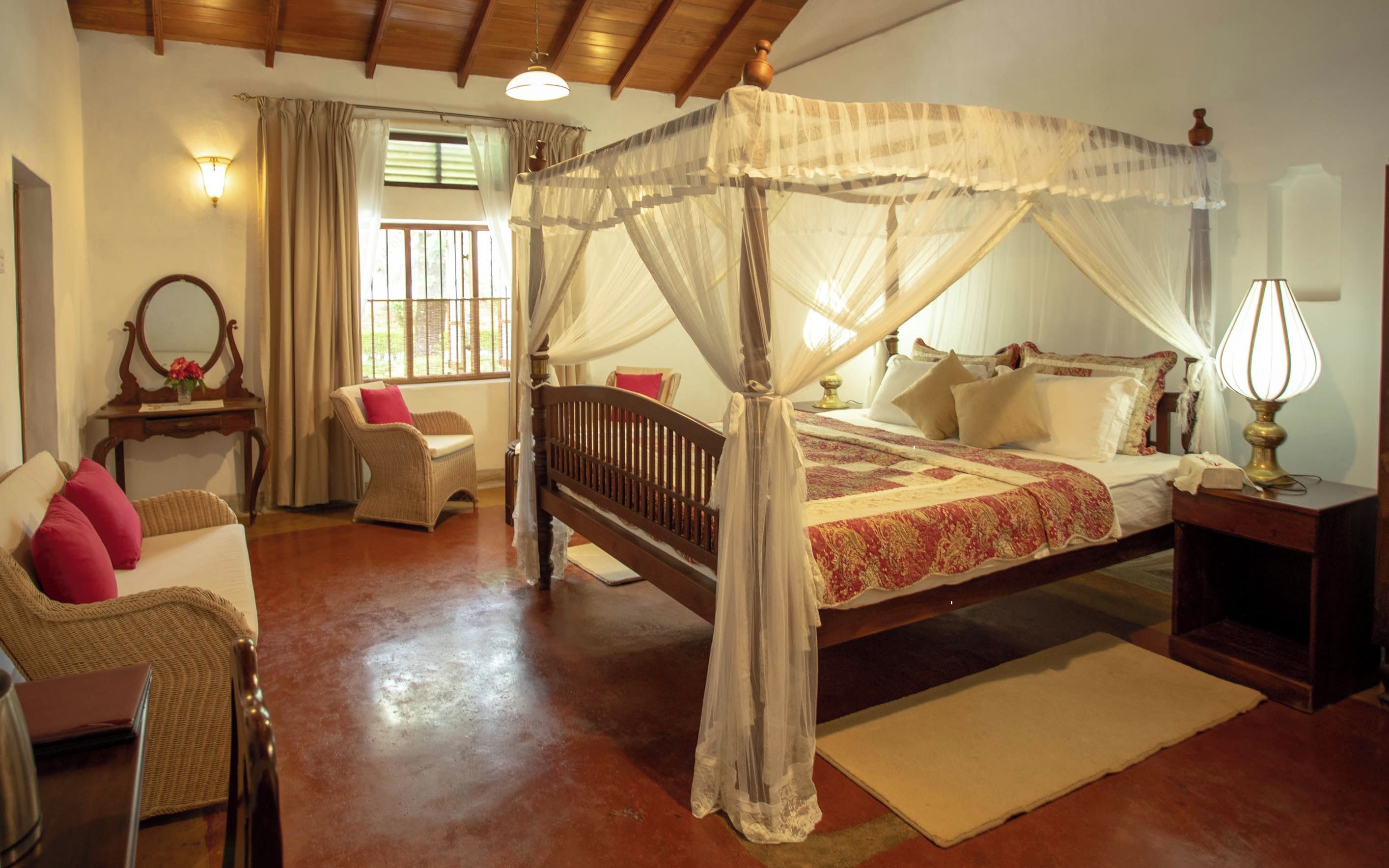 Un dormitorio con cama con dosel, un rincón de descanso con una silla y una mesa, y una ventana con cortinas.