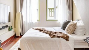 Een slaapkamer met een bed, kussens, een deken, een televisie en een raam met gordijnen.