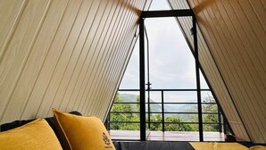 Een chalet met schuin dak, twee bedden met gele kussens en uitzicht op groen en bergen door grote ramen.