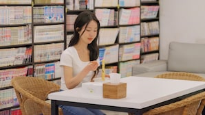 Eine Person sitzt an einem Tisch mit einem Buch, einer Tasse und einer Kiste vor einem Bücherregal voller Bücher.