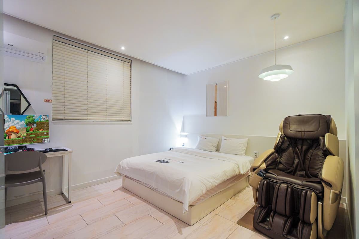 Un dormitorio con cama, escritorio con ordenador, sillón de masaje y ventana con persianas.