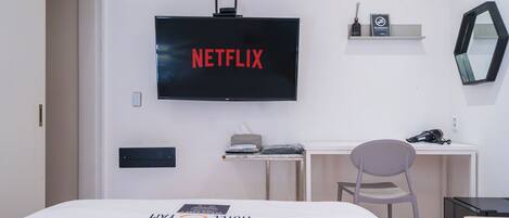 A hotel room with a bed, a desk with a chair, a TV mounted on the wall, and a small shelf with a sign.