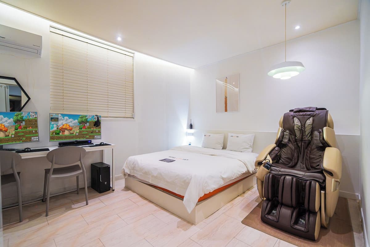 Un dormitorio con cama, escritorio, silla, sillón de masaje y aire acondicionado instalado en la pared.