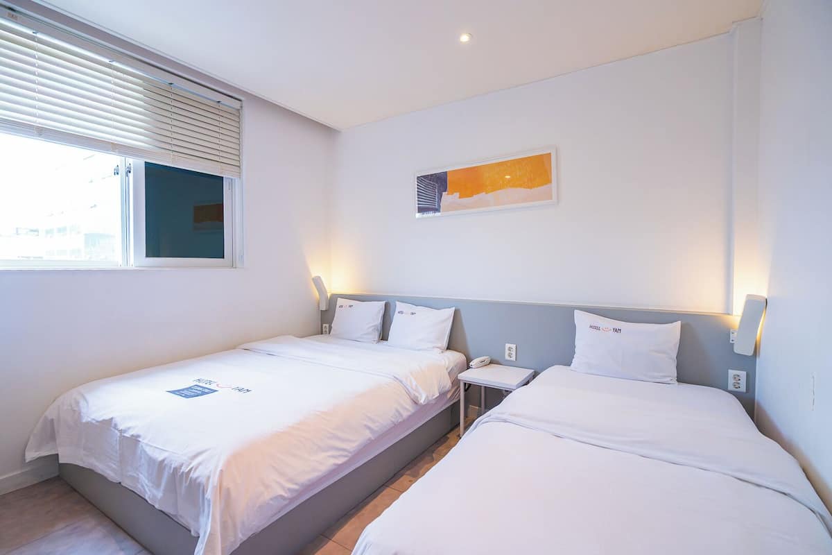 Habitación de hotel con dos camas, una mesita de noche con teléfono, una ventana con persianas y una lámpara fijada a la pared.