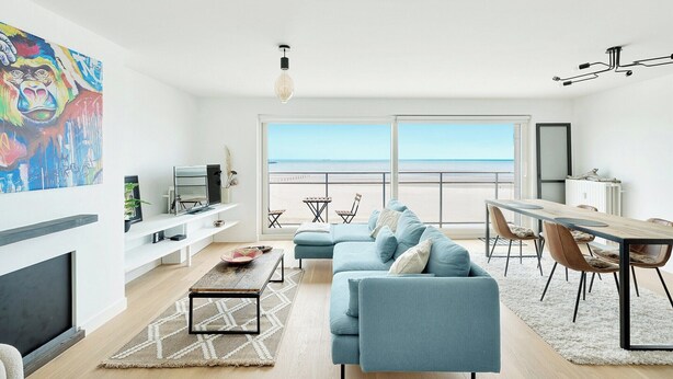A modern living room with a large window, a sofa, a dining table, and a TV. - Renovated apartment in Knokke-Heist with views (HEIST AAN ZEE)