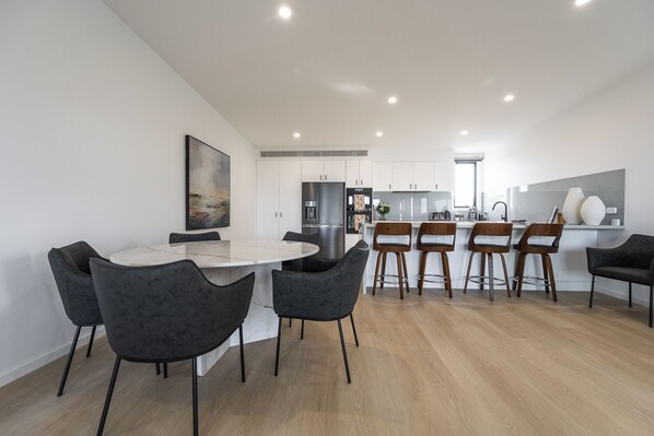 Una cocina moderna con armarios blancos, un refrigerador de acero inoxidable y un comedor con una mesa de mármol y sillas negras.